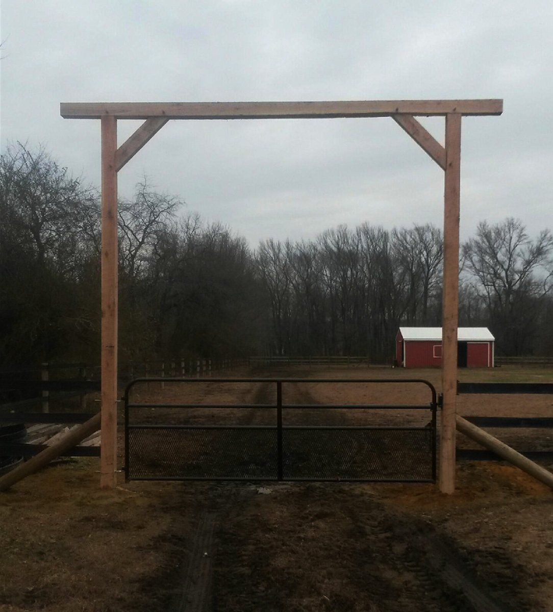 Wooden gate frame over a fenced dirt path, with trees and a small red barn in the background.
