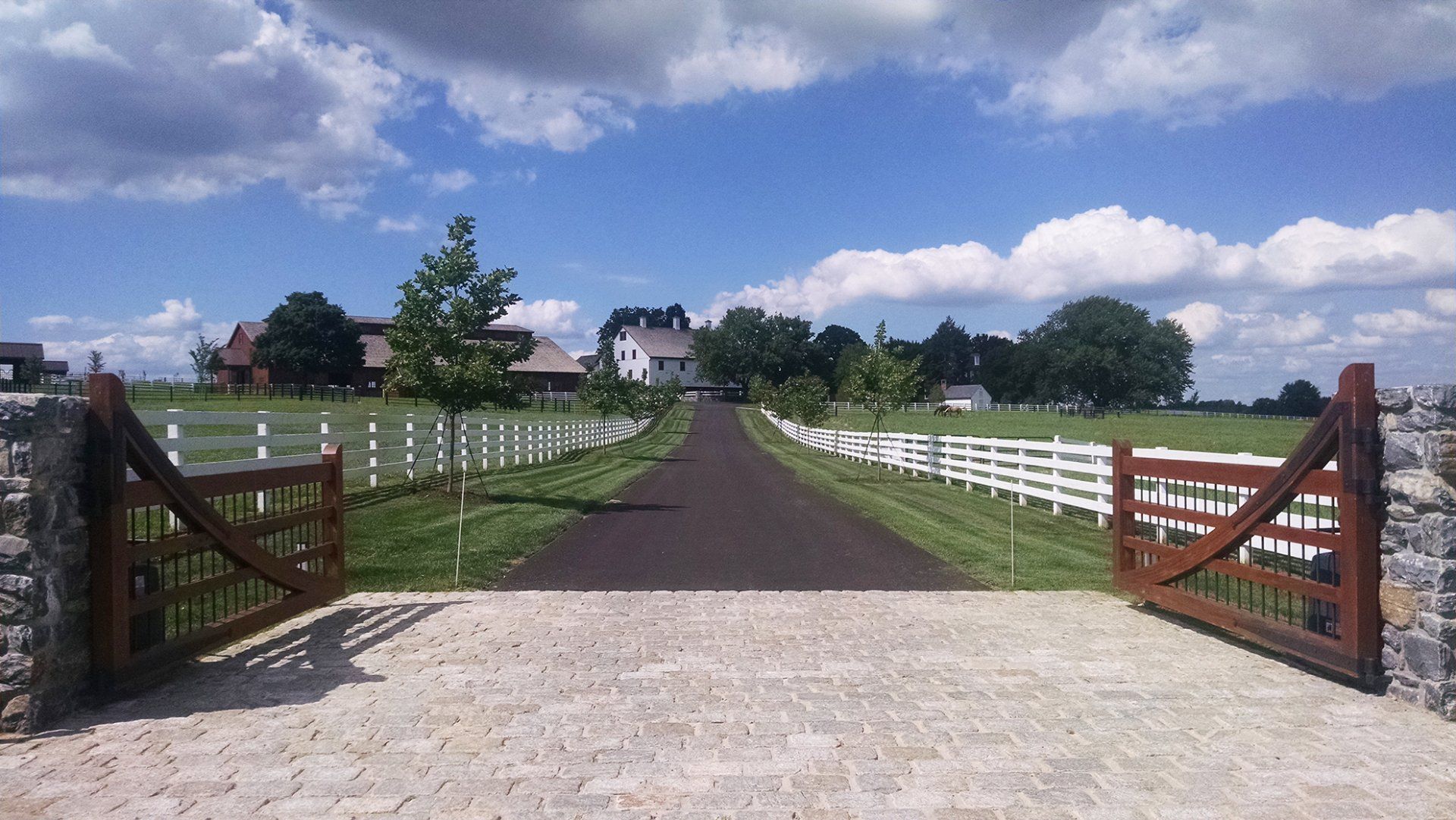 Open farm gate leading to a gravel driveway through a green field under a blue sky with clouds