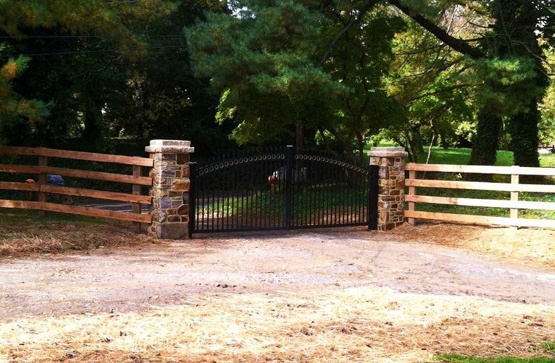 Black iron gate between brick pillars at a wooded driveway entrance.
