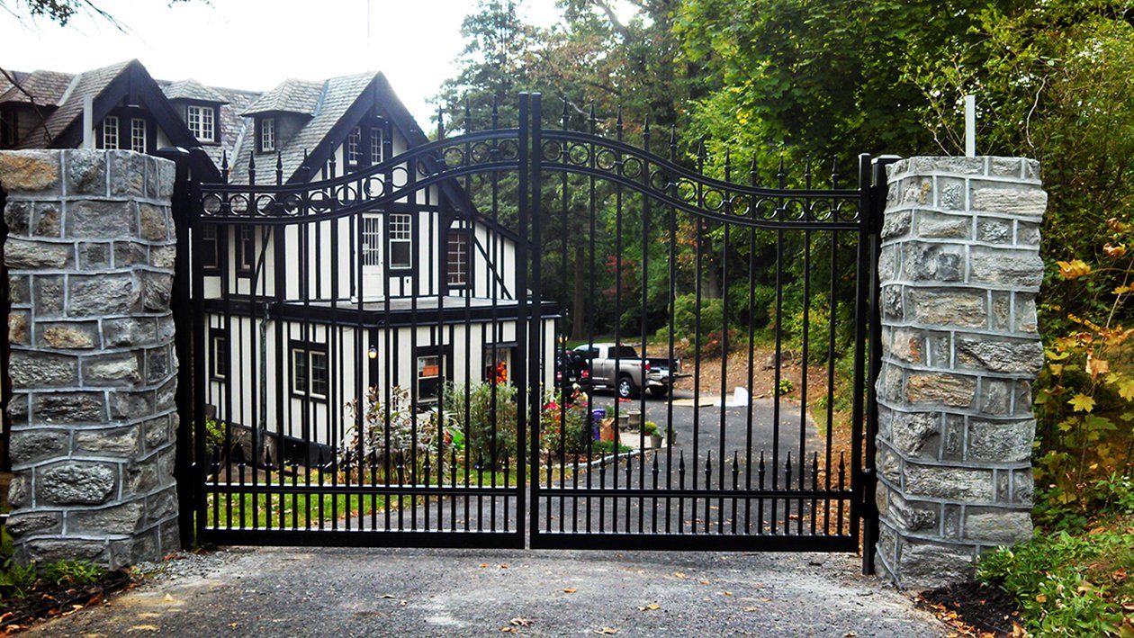 Black wrought-iron driveway gate between stone pillars in front of a house