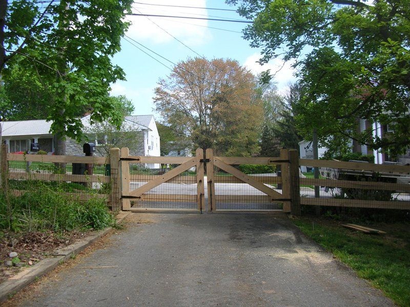 Wooden farm gate closing a paved driveway, with trees and fences on both sides.