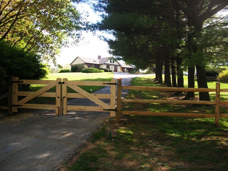 Wooden gate and fence across a driveway leading to a house in a sunny rural yard.