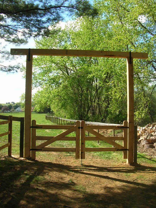 Wooden farm gate under a simple frame, opening to a sunlit dirt path and trees beyond.