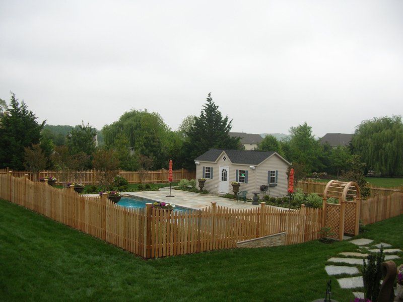 Fenced backyard play area with a small playhouse, sandbox, and toy structures on a grassy lawn