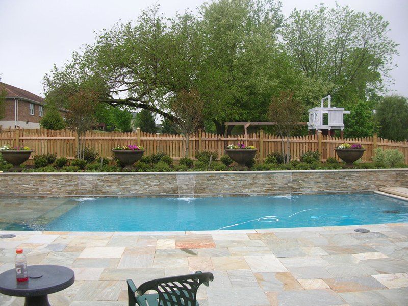 Backyard pool with stone patio, chairs, fence, and lush trees behind it