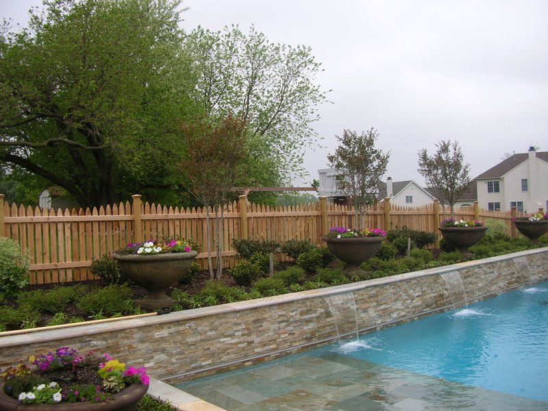 Backyard pool with stone edging, flower beds, wooden fence, and houses beyond under a cloudy sky