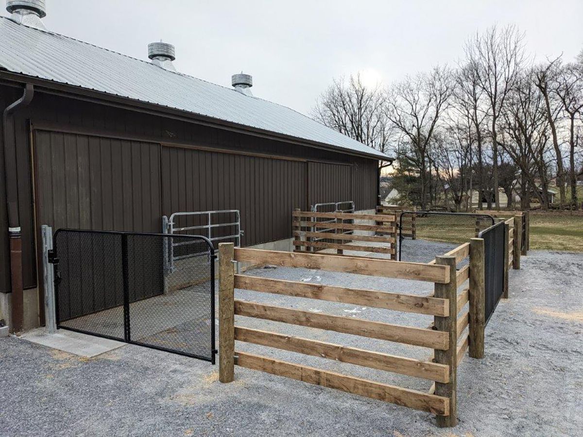 Gravel yard beside a dark barn with wooden fence panels and metal gates under a cloudy sky
