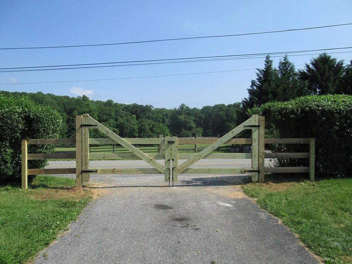 Wooden farm gate blocking a gravel driveway, with green hedges and fields under a blue sky