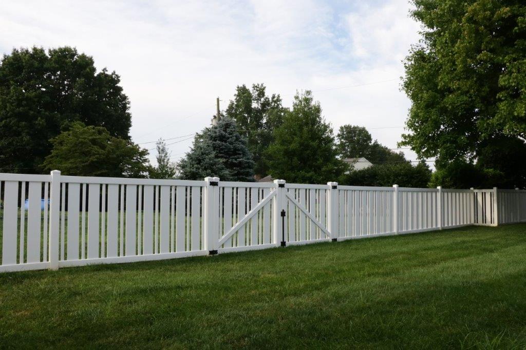White wooden fence with a gate along a grassy yard, with trees and sky in the background