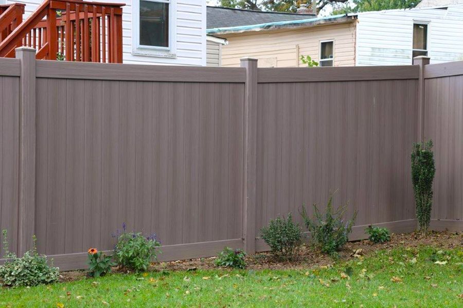 Gray privacy fence in a backyard with grass, shrubs, and a deck visible above it