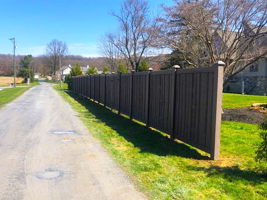 Gravel road beside a long black wooden fence with green grass and trees on a sunny day
