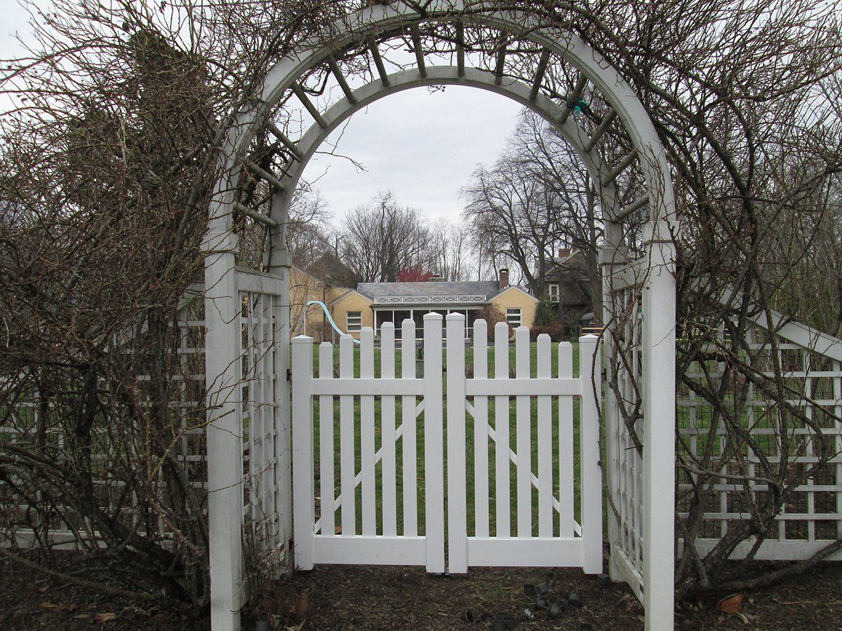 White picket gate under a vine-covered stone arch at the entrance to a garden path