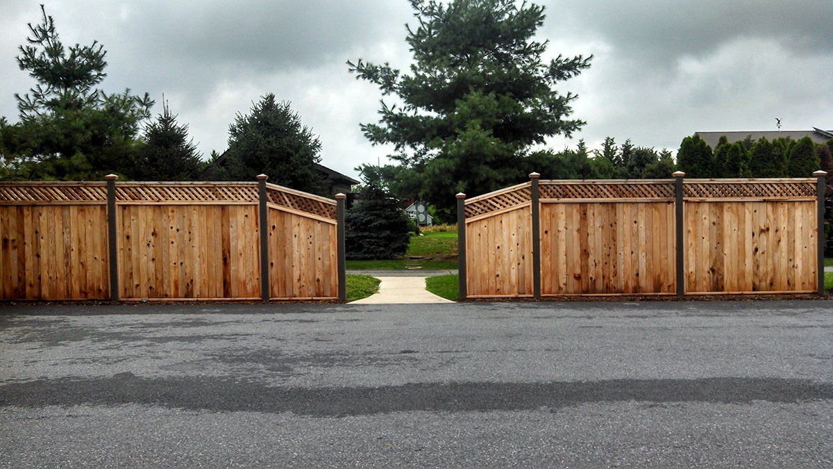 Wooden fence gates framing a path opening to trees and a house beyond a paved road