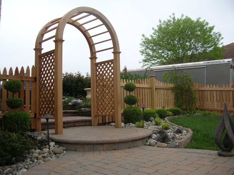 Wooden garden arbor over a brick patio, surrounded by trimmed shrubs and a picket fence.