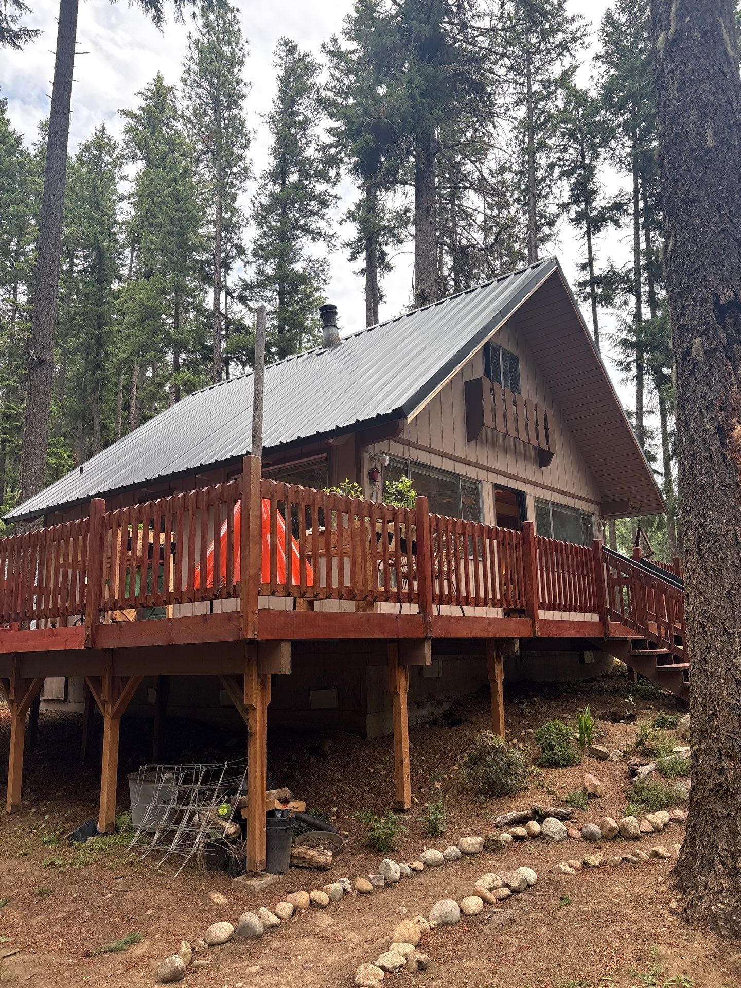 Rustic cabin on stilts with a gray roof and orange deck railing in a pine forest