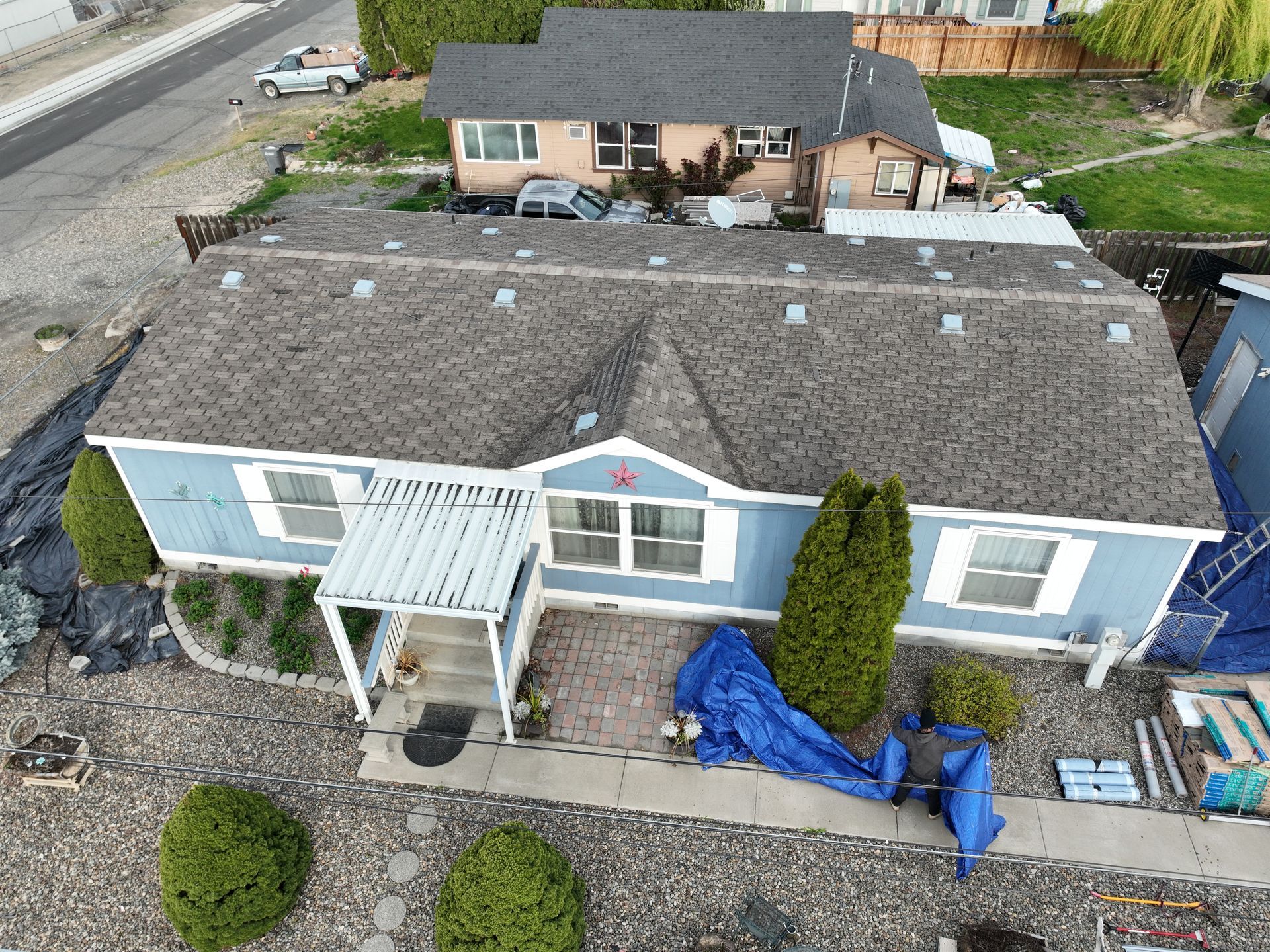 Aerial view of a blue house with a gray roof, porch, and yard covered with tarps and materials.