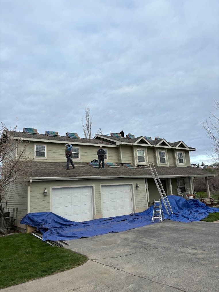 Workers on a roof with blue tarps and ladders outside a two-story house with three garage doors