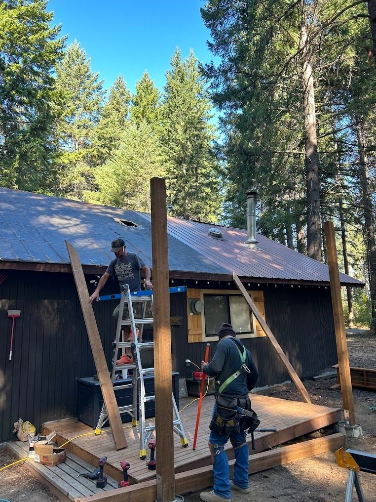 Two workers on ladders repairing a cabin porch roof in a wooded area