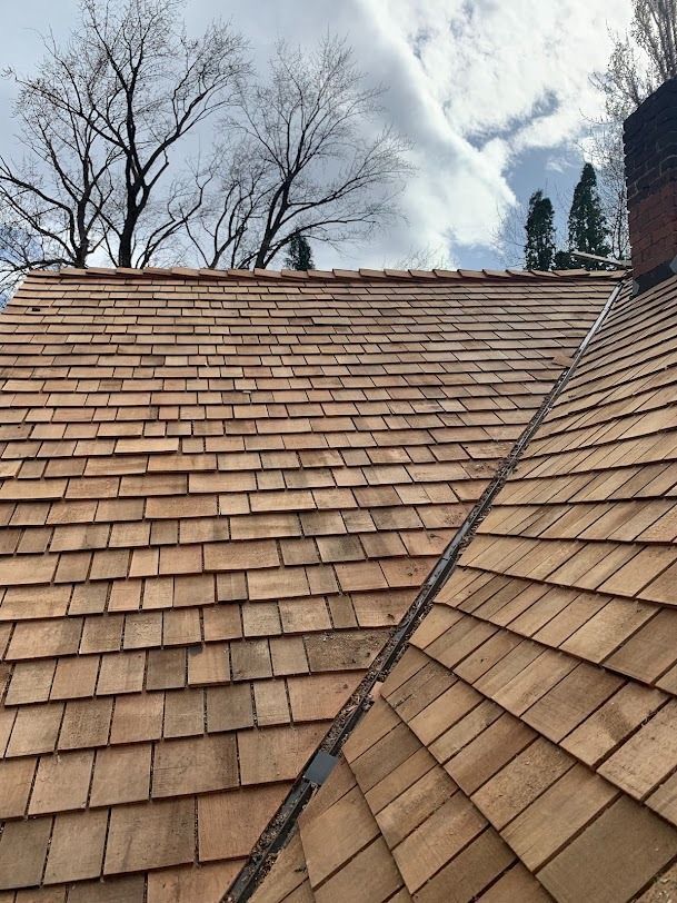 Tan shingled roof with a valley seam beneath leafless trees and cloudy sky