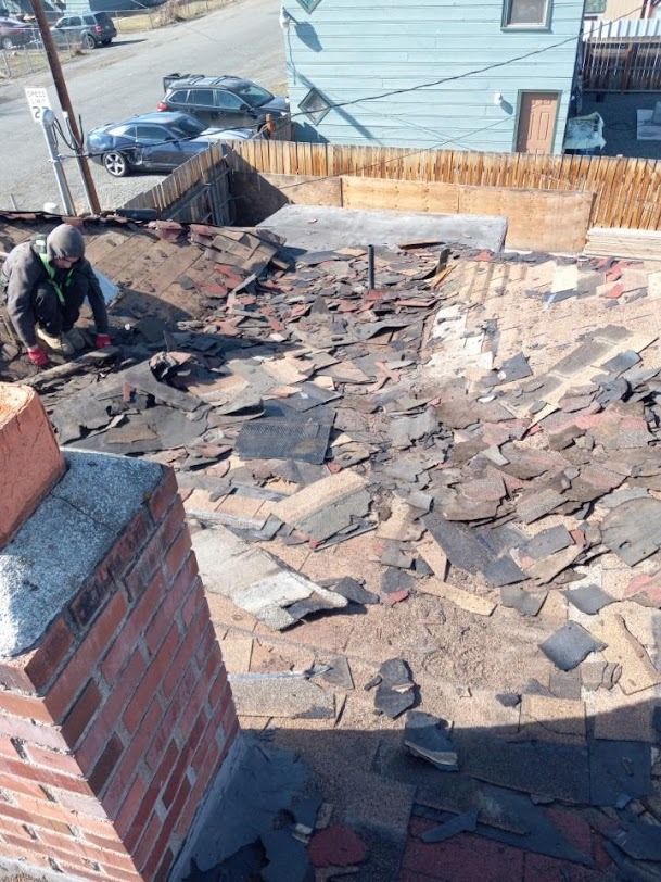 Worker repairing a sloped shingle roof beside a brick chimney on a sunny day