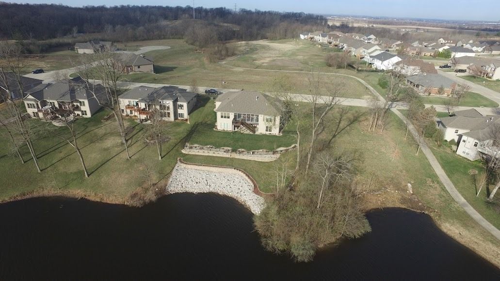 Aerial view of lakeside homes, lawns, and roads beside a wooded shoreline