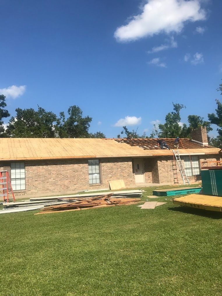 Partially renovated brick house with exposed roof, lumber stacks, and green lawn under a blue sky