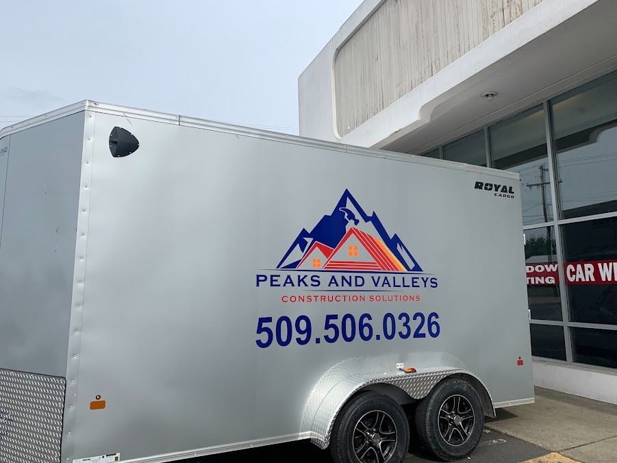 White Peaks and Valleys cargo trailer with blue logo and phone number parked by a building.