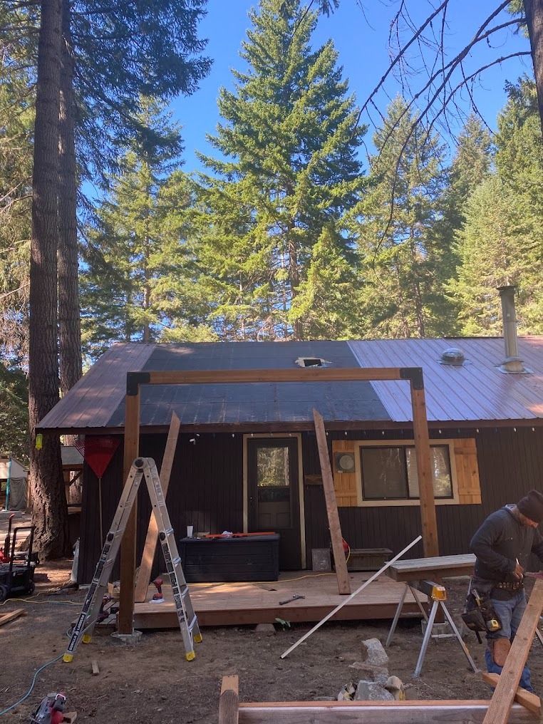 Cabin porch under construction with workers, wooden A-frame, and tall pine trees in the background