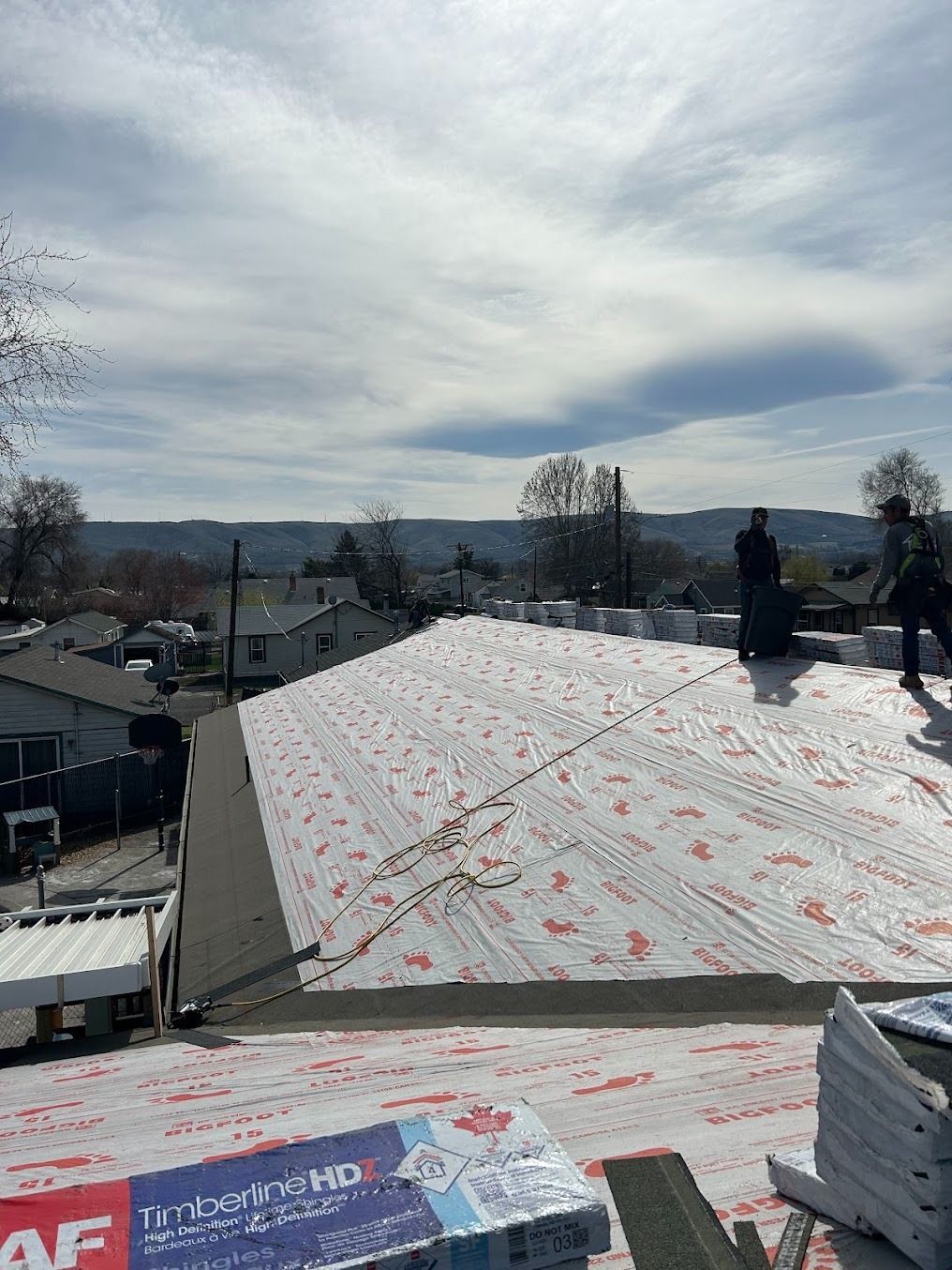 Roofer on a shingled roof under cloudy sky, with stacks of roofing materials in the foreground.