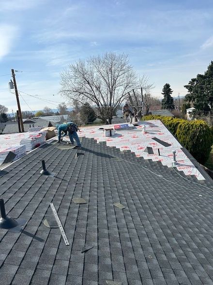 Workers installing roofing underlayment on a gray roof, with white sheets and a bare tree in the background
