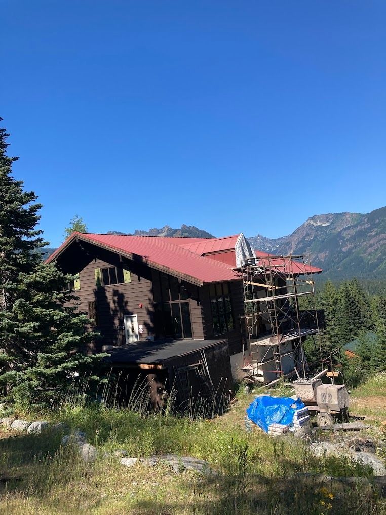 Mountain cabin with a red roof under scaffolding, surrounded by trees and mountains under a clear blue sky
