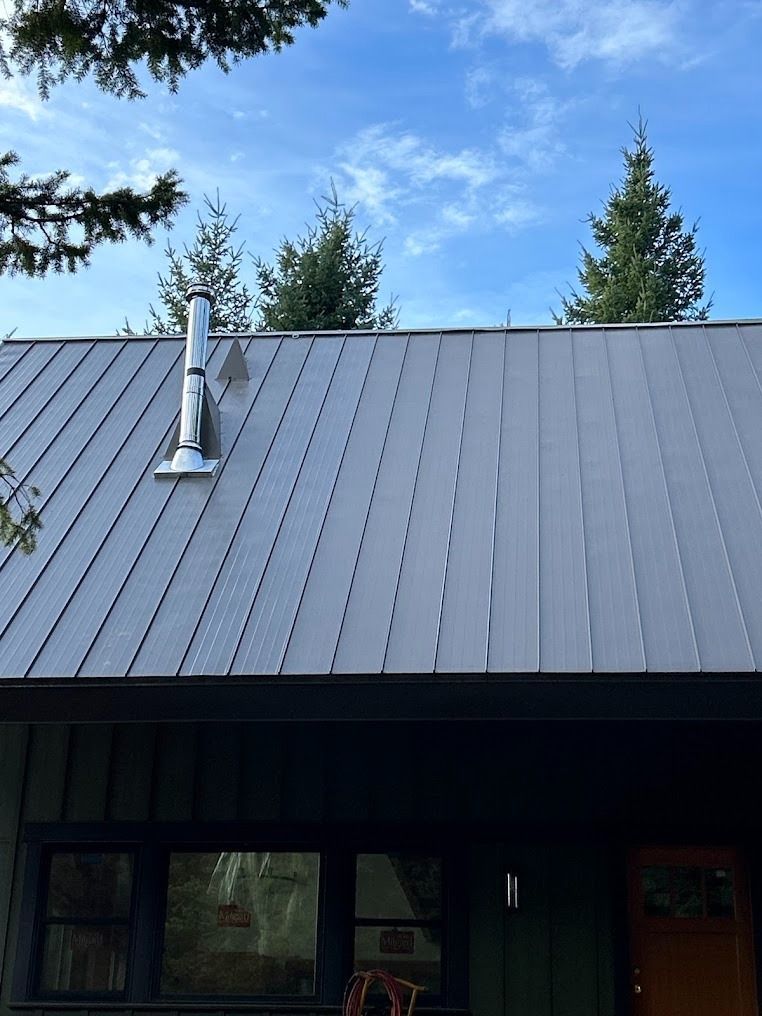 Dark metal roof on a cabin with a silver chimney under a blue sky.
