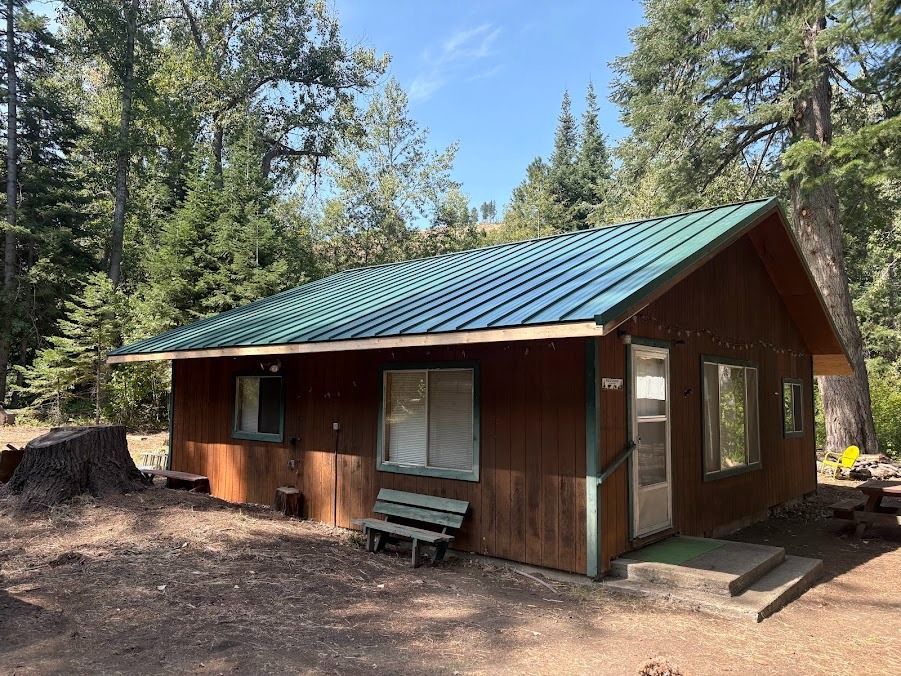 Small brown cabin with a green metal roof in a forest clearing, with a bench out front.