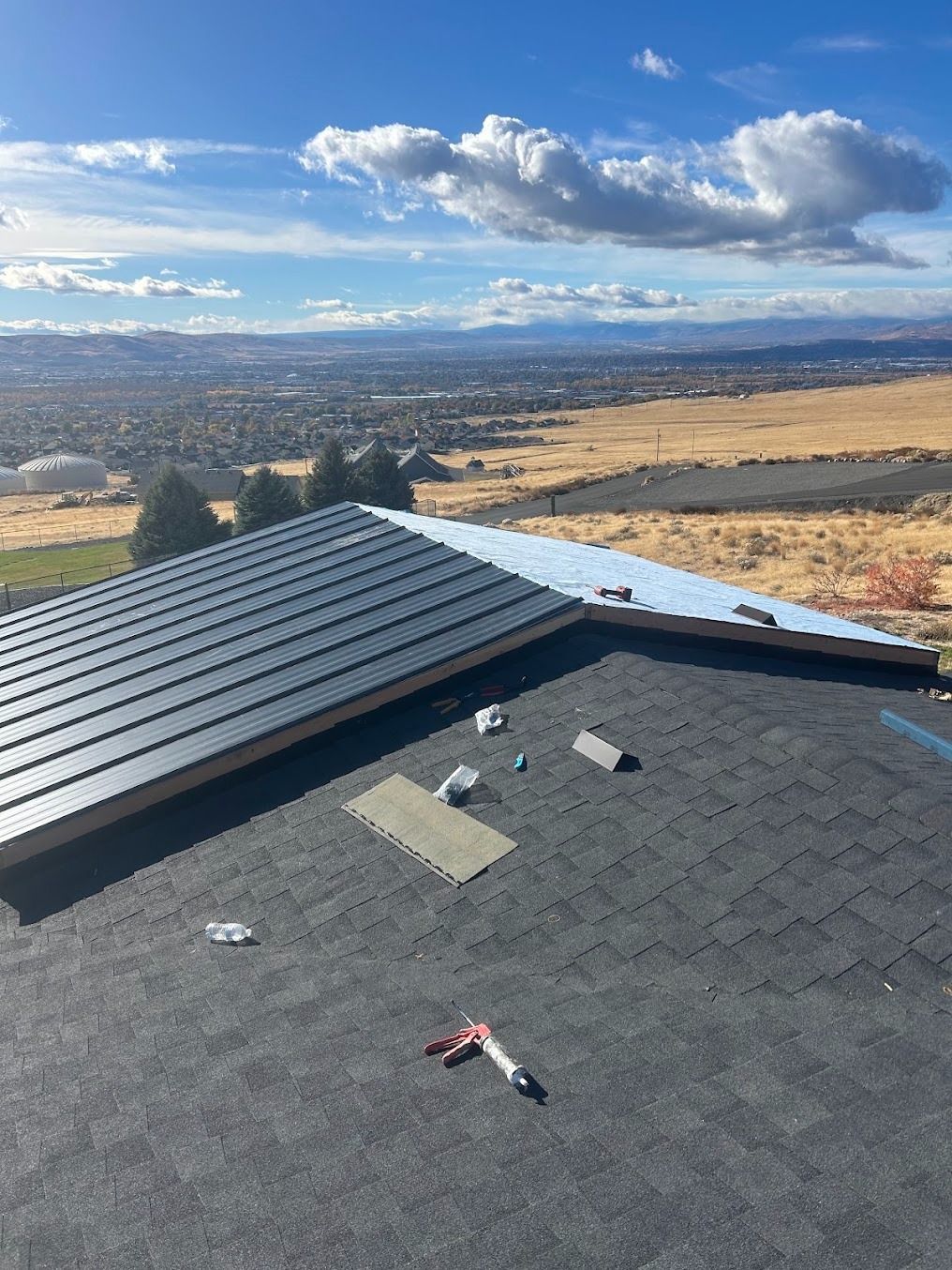 Rooftop with shingles and patchwork repairs overlooking a wide valley under a cloudy blue sky
