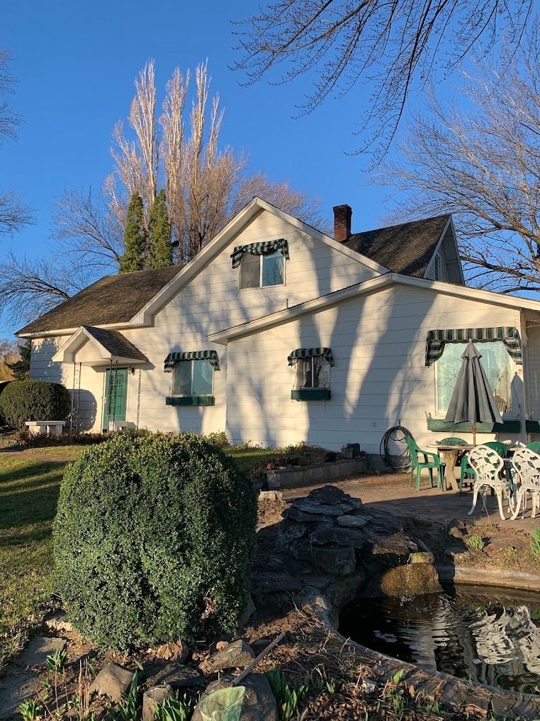 Cream-colored cottage with green shutters, patio furniture, and a pond under a clear blue sky