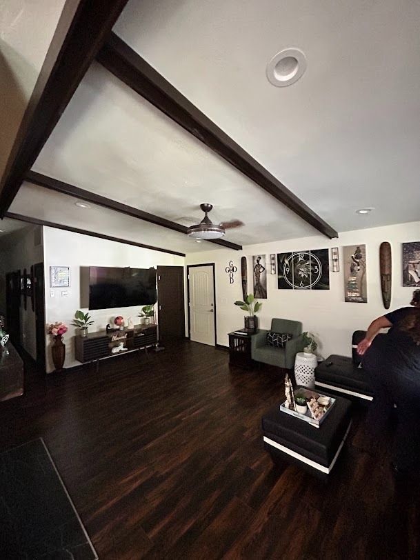 Modern living room with dark wood beams, TV, coffee table, plants, and a seated person on a sofa