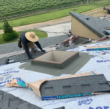 Worker installing a skylight on a shingled roof, with roofing materials and a rural yard below.