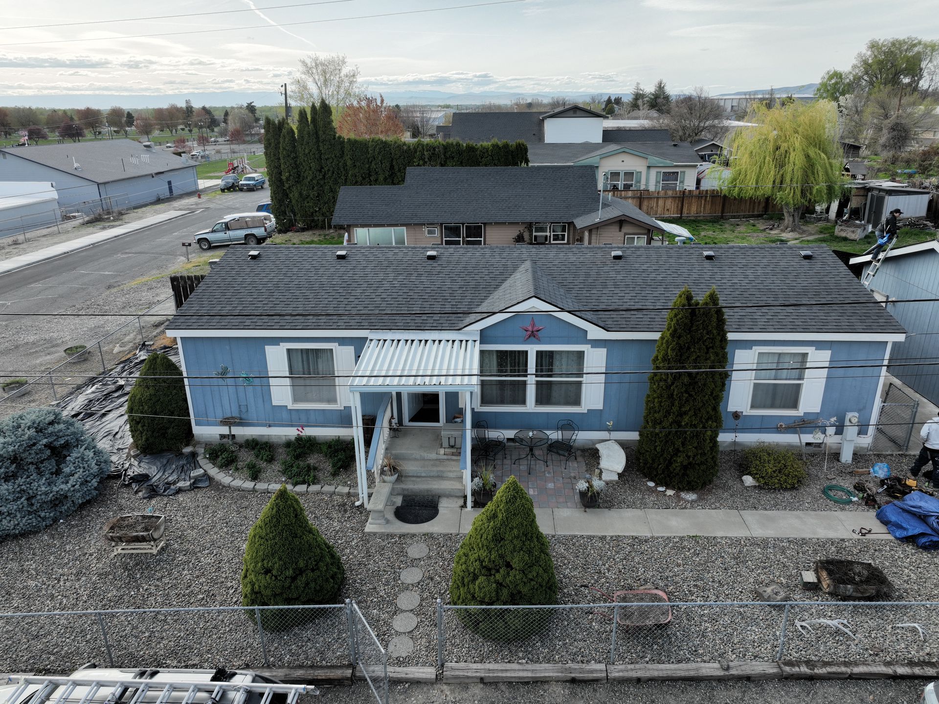 Aerial view of a blue house with a dark roof and front yard, surrounded by a fenced landscape.