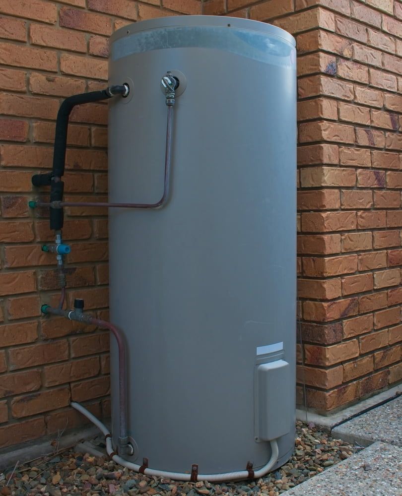 Grey Outdoor Water Heater Tank Beside a Brick Wall With Exposed Pipes and Gravel Base — JAJ Plumbing in Mount Austin, NSW