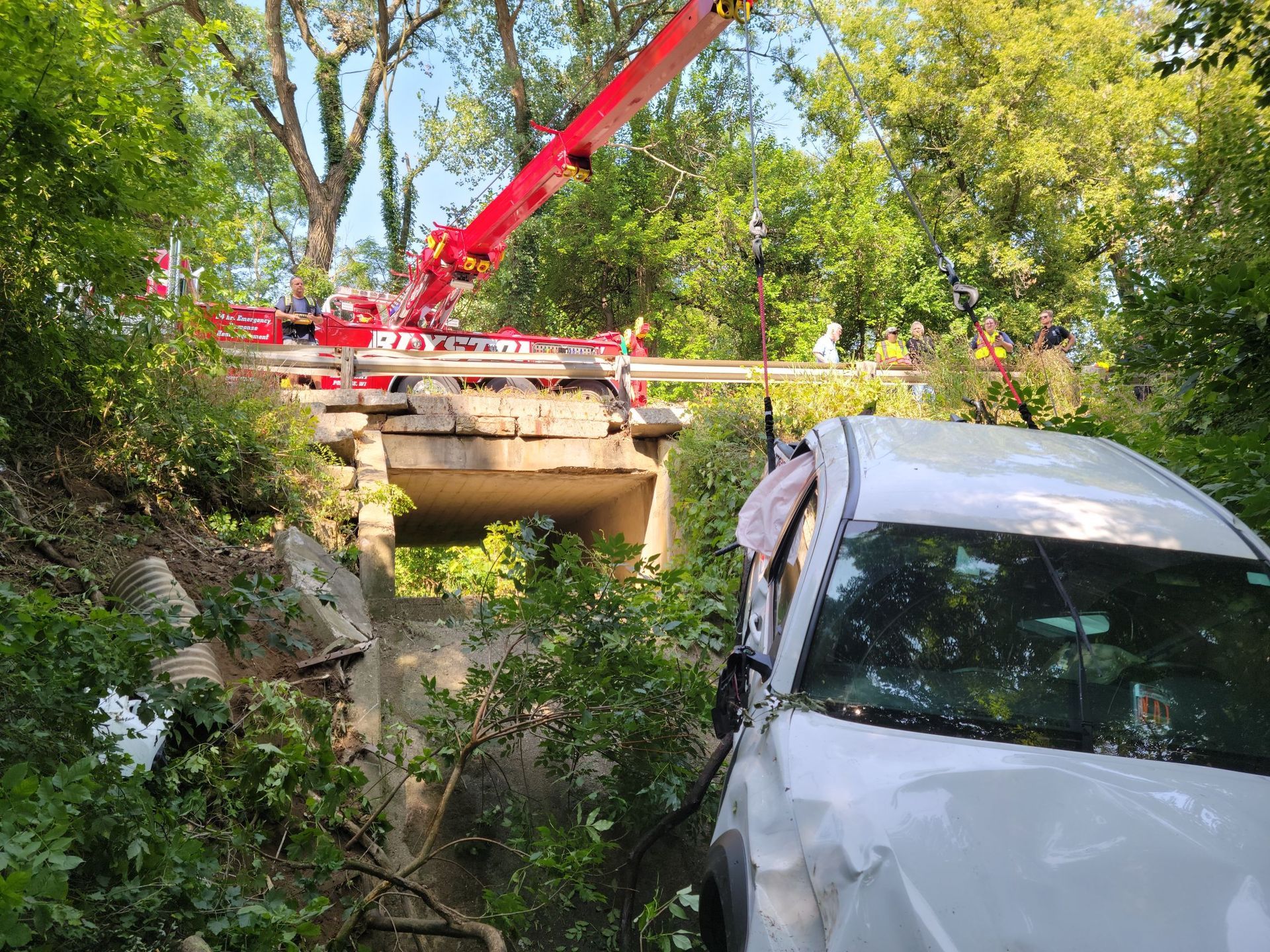White car crashed under a damaged bridge; red crane overhead. Trees surround the scene.