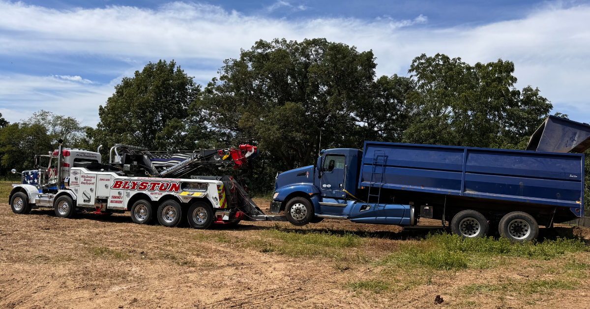 Tow truck pulling a blue dump truck on a dirt road, trees in background.