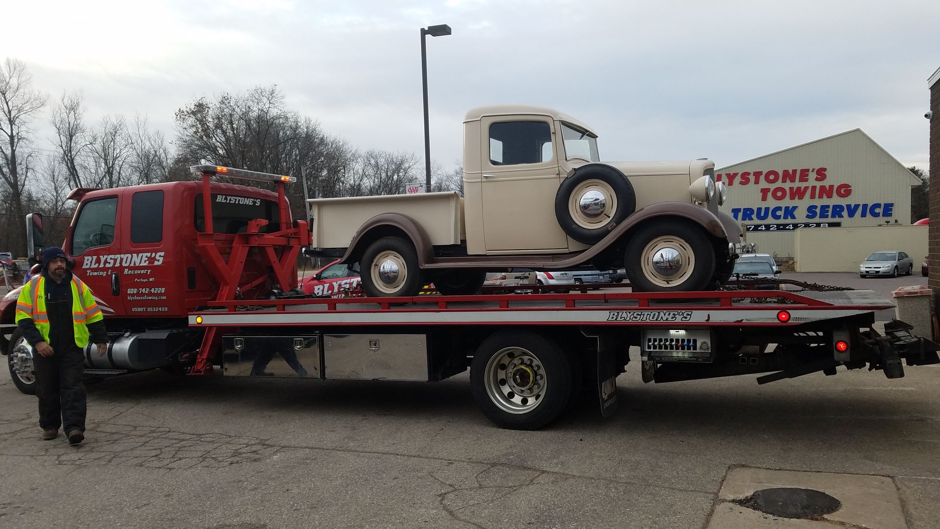 A vintage beige pickup truck is towed on a red flatbed truck in front of a towing service building.