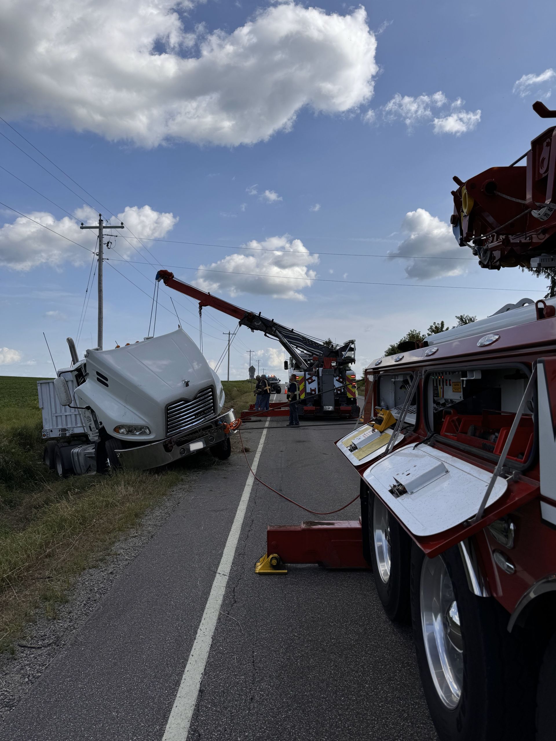 Tractor-trailer on side of road being towed by two tow trucks, with a blue sky and white clouds.