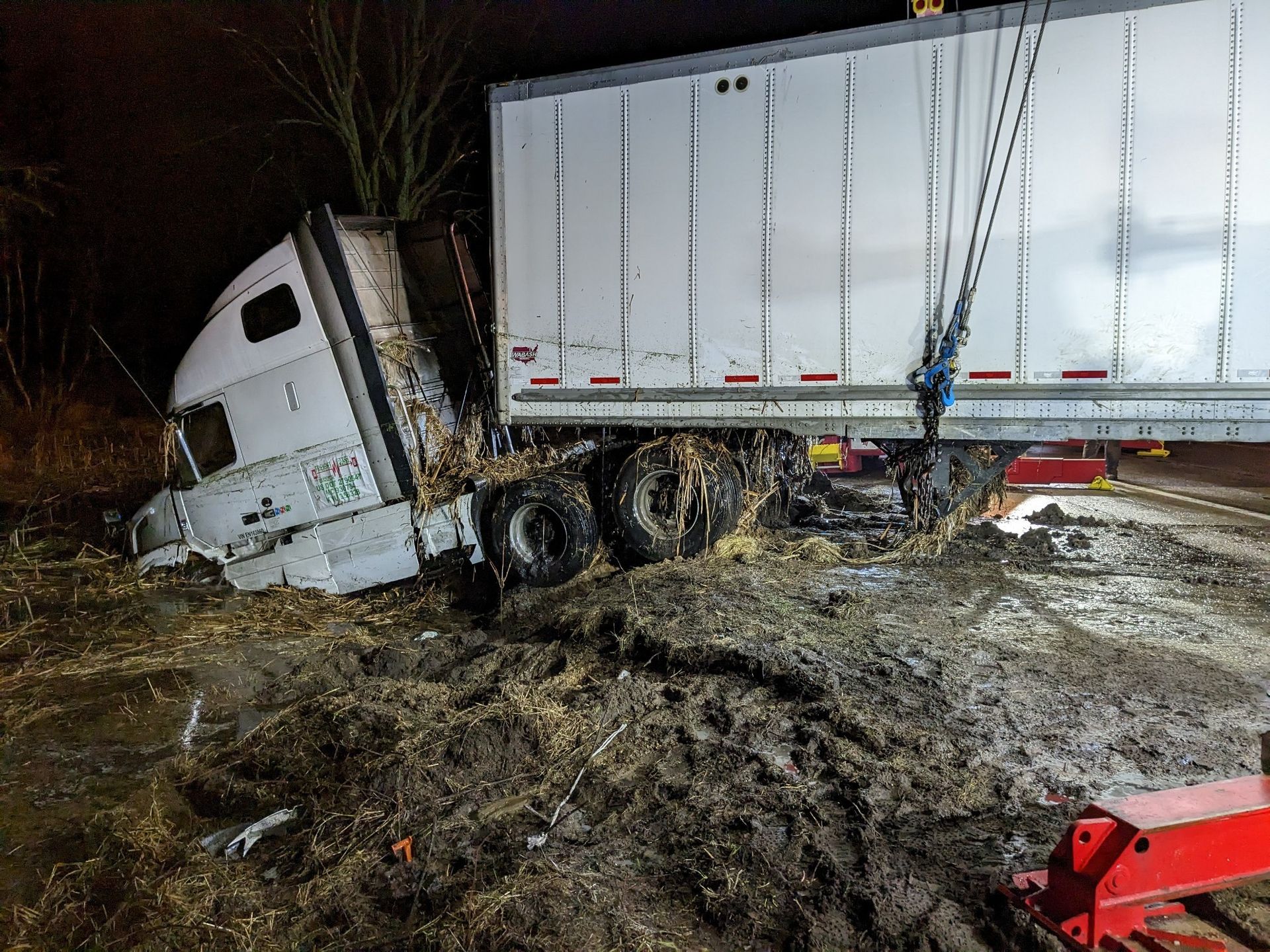 Semi-truck stuck in mud, cab tilted. Trailer being pulled by tow truck. Night scene, dark.