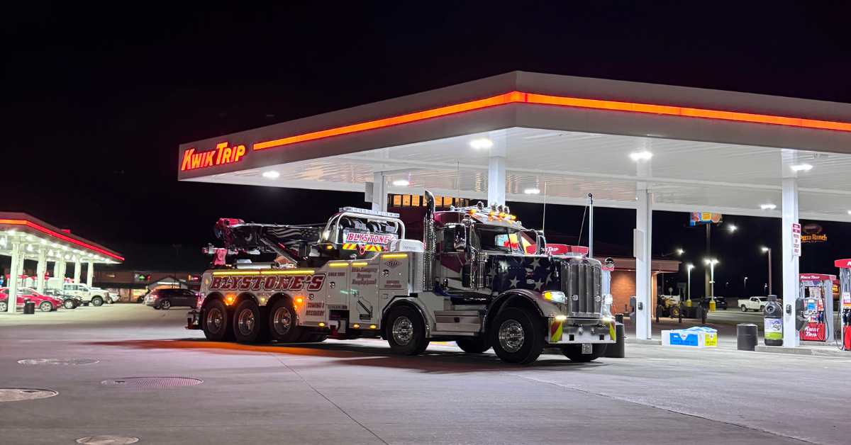 A tow truck parked at a gas station at night. The gas station has bright lights.