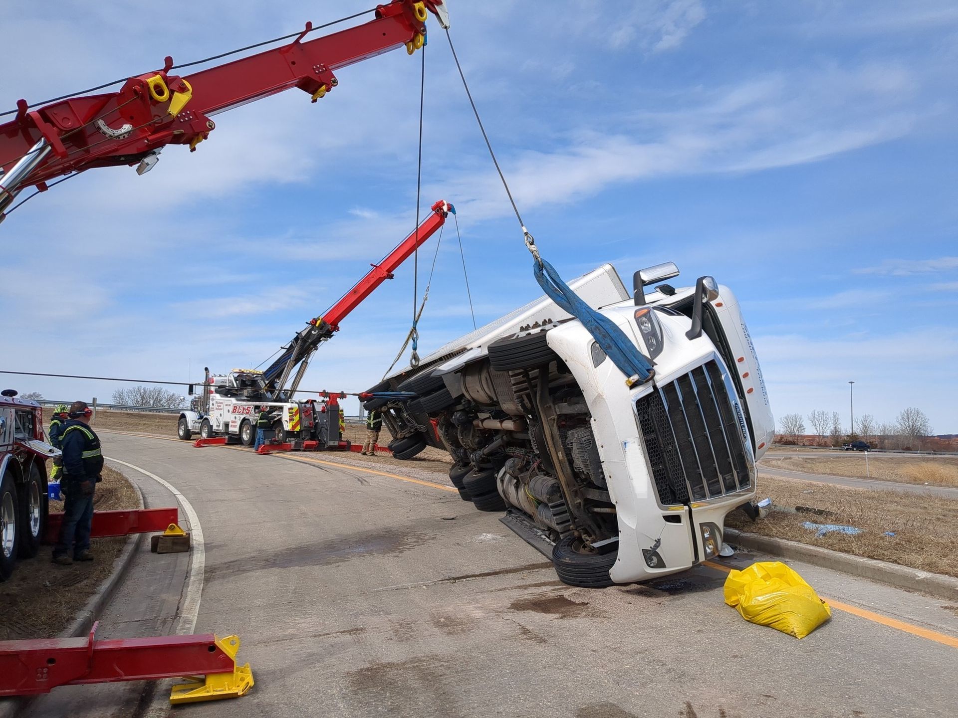 A semi-truck overturned on a road being lifted by two tow trucks, blue sky, yellow caution bags.