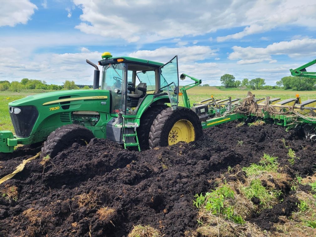 Green John Deere tractor stuck in muddy soil while plowing a field under a partly cloudy sky.