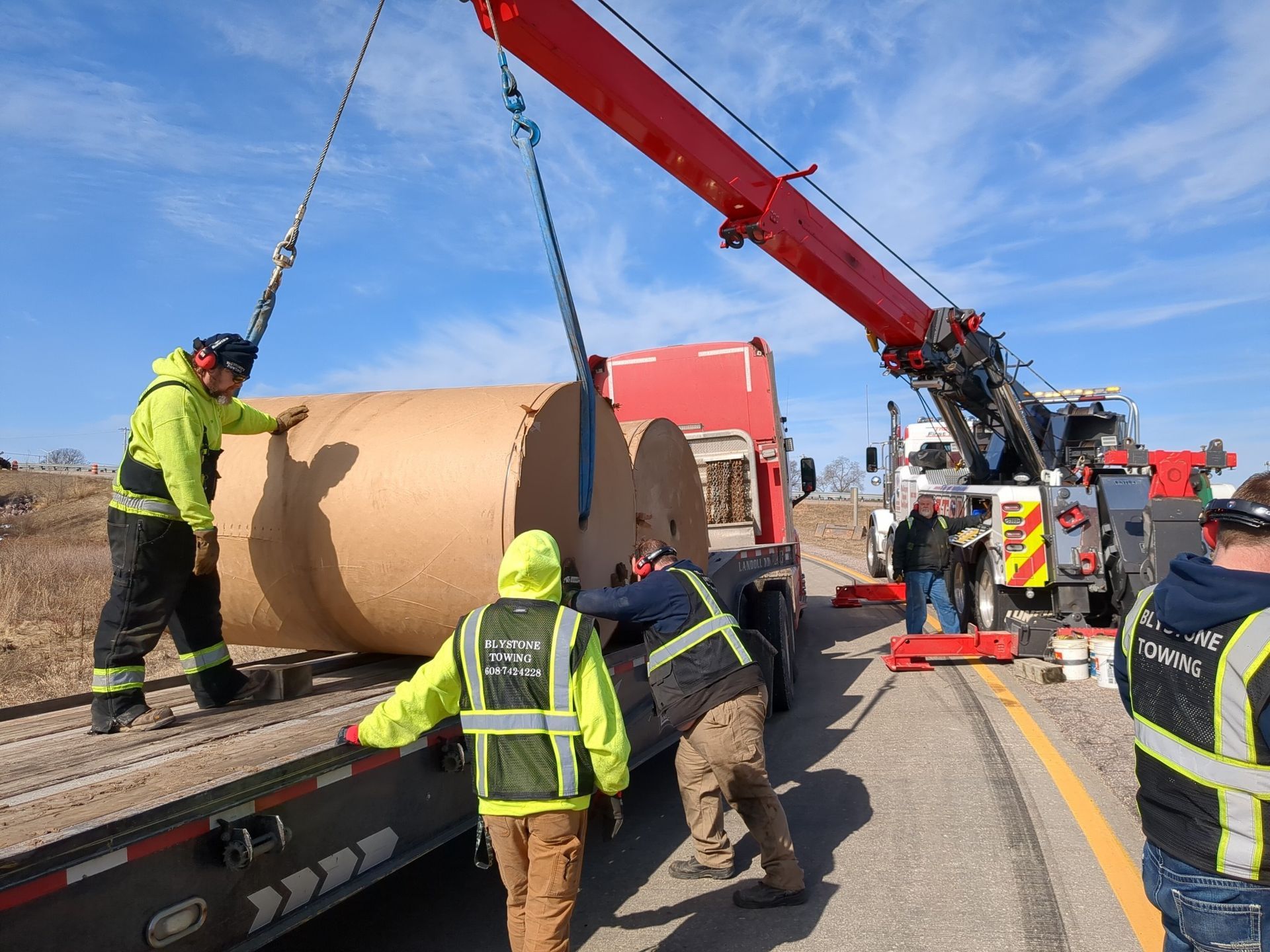 Workers using a crane to unload large paper rolls from a flatbed truck on a highway.