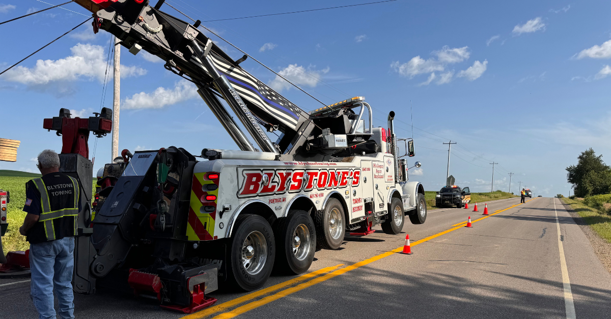 Tow truck on a rural road, removing a vehicle. Safety cones and officer present. Bright sky and green fields.