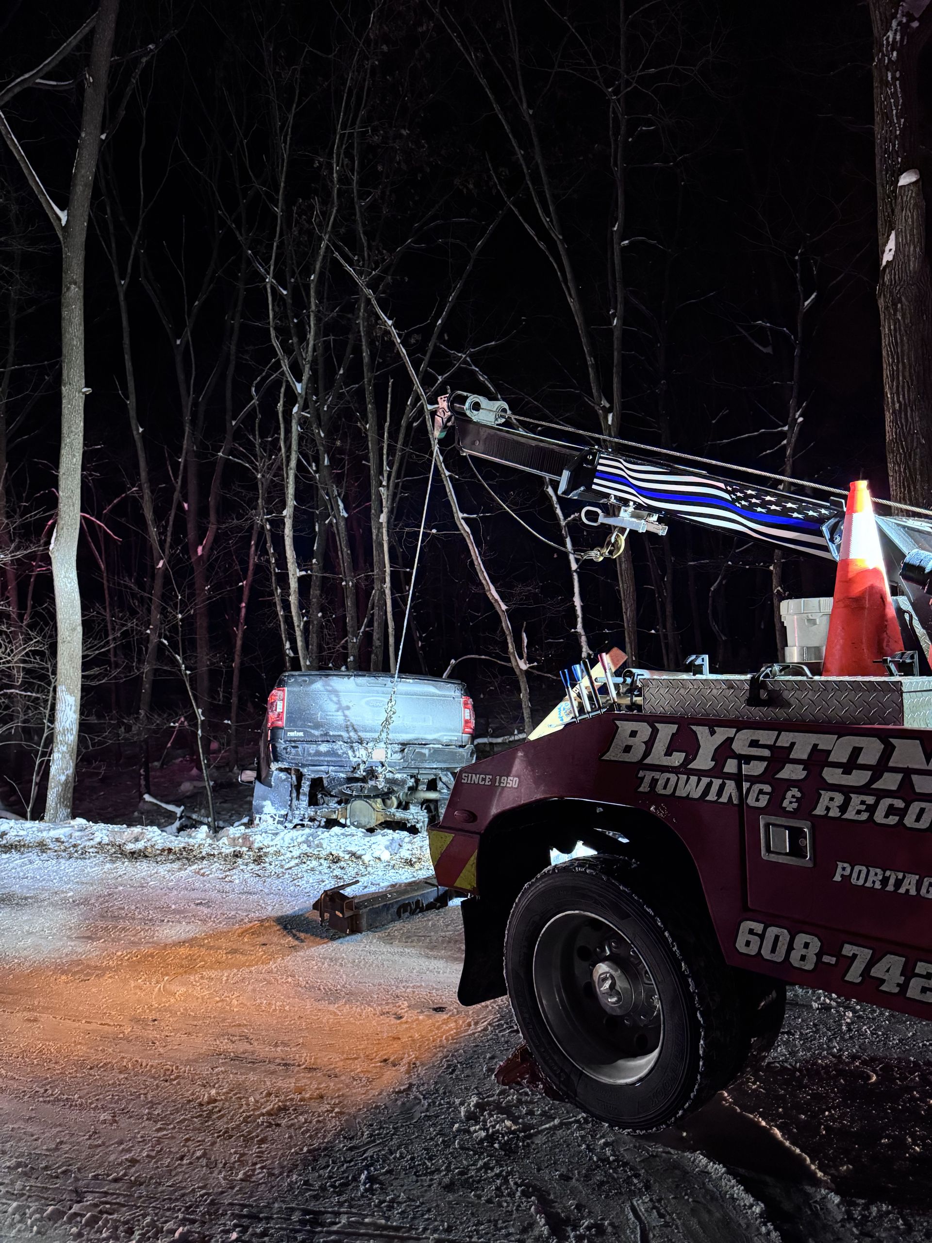 Tow truck at night, recovering a car that crashed into trees. Snowy scene, orange cone on the truck.